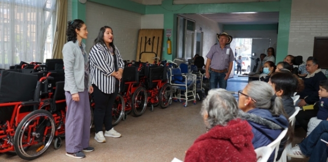 Dos mujeres hablan frente a un grupo de personas sentadas en un salón comunitario, mientras varias sillas de ruedas y equipos de movilidad están organizados al fondo del espacio.